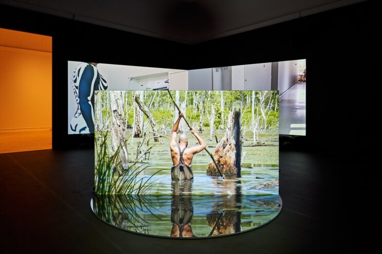 <p>A.K. Burns (U.S., b. 1975). <em>What is Perverse is Liquid (NS 0000)</em>. 2023. 3-channel HD video installation (color, 5.1 sound), 35:54 mins. Two projection screens, free-standing projection wall, plexiglass, sandbags, sand, rubber pool liner, and pennies. Courtesy of the artist. [Installation view, <em>A.K. Burns: What is Perverse is Liquid,</em> Henry Art Gallery, University of Washington, Seattle. 2024]. Photo: Jonathan Vanderweit</p>
